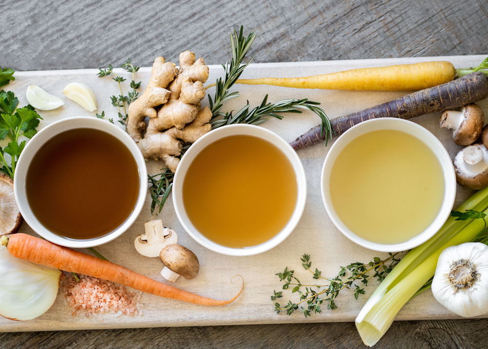 Three bowls of different colored broth centered on a cutting board surrounded by fresh vegetables and herbs